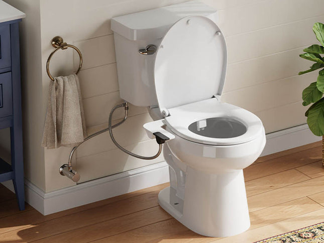 A white toilet with the lid open, shown installed in a bathroom with wood flooring, a beige paneled wall, a blue cabinet, a towel ring with a beige towel, and a green plant.