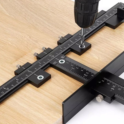 A black metal square drill guide with ruler markings and adjustment knobs, shown in use with a drill press against a light wood surface.