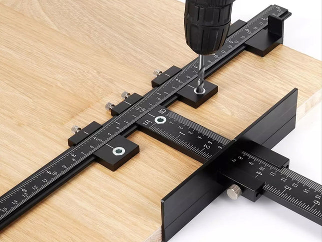 A black metal square drill guide with ruler markings and adjustment knobs, shown in use with a drill press against a light wood surface.