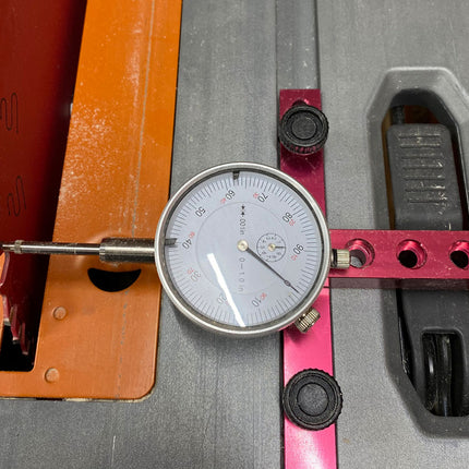 A dial caliper or precision measuring tool with a round analog gauge, silver probe, and red adjustment arm, shown resting on a gray metal surface next to orange and pink guide rails.