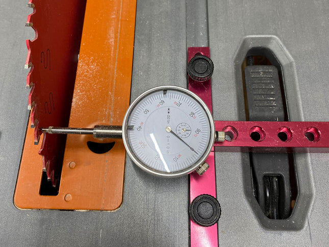 A dial caliper or precision measuring tool with a round analog gauge, silver probe, and red adjustment arm, shown resting on a gray metal surface next to orange and pink guide rails.