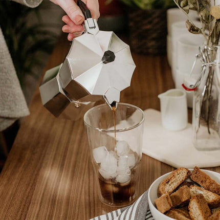 Person pouring coffee from a Moka pot into a glass with ice on a wooden table.