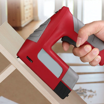 A red and gray cordless nail gun being held by a hand while driving a nail into a light wood frame, with a softly blurred indoor background.