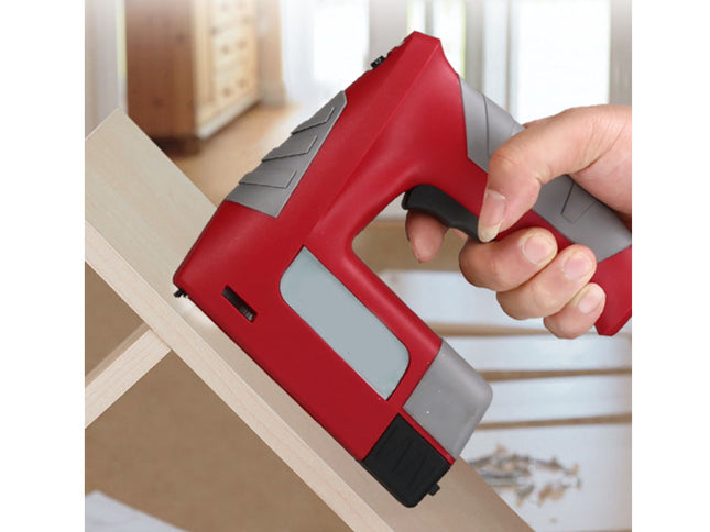A red and gray cordless nail gun being held by a hand while driving a nail into a light wood frame, with a softly blurred indoor background.