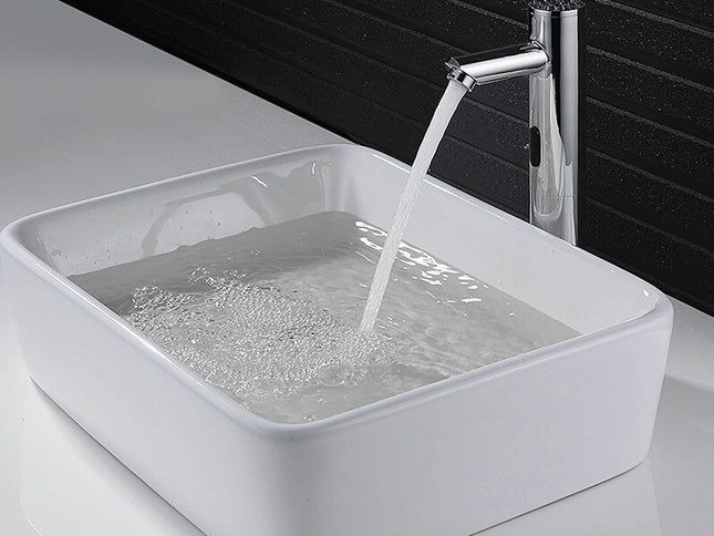 A modern rectangular white ceramic vessel sink with running water from a tall chrome faucet against a dark tiled wall.