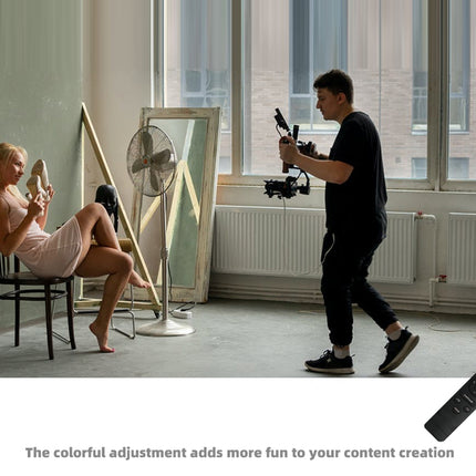 A lifestyle scene in a studio showing a woman sitting on a chair holding a phone while a man walks with a camera rig. Two tall vertical light panels, a floor fan, and a mirror are visible in the room.