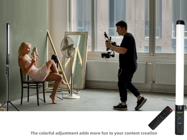 A lifestyle scene in a studio showing a woman sitting on a chair holding a phone while a man walks with a camera rig. Two tall vertical light panels, a floor fan, and a mirror are visible in the room.