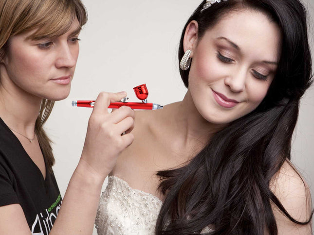 A woman with long dark hair and a tiara receives a makeup application from another woman holding a red airbrush pen. The woman on the right is wearing a white lace dress and silver earrings against a plain light background.