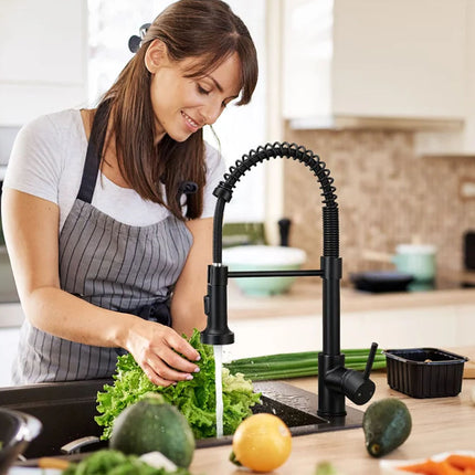 A woman in a kitchen wearing a gray apron stands at a sink and washes leafy greens under a modern black pull-down faucet. Fresh vegetables, including avocados, a lemon, and carrots, are visible on the counter.