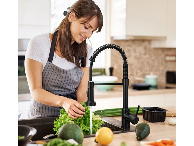 A woman in a kitchen wearing a gray apron stands at a sink and washes leafy greens under a modern black pull-down faucet. Fresh vegetables, including avocados, a lemon, and carrots, are visible on the counter.