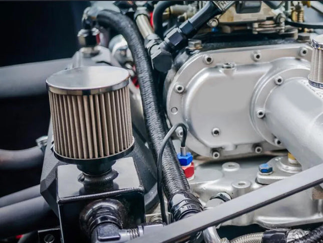 Close-up of a car engine with a silver metal intake manifold, black hoses, and a cylindrical air filter on a black mounting block.