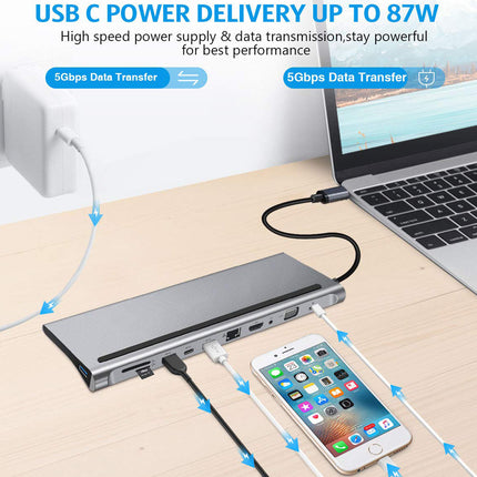 A silver USB-C hub shown on a light wood desk, connected to a wall charger, a laptop, and a smartphone. Blue text and arrows highlight USB-C power delivery up to 87W, 5Gbps data transfer, and high-speed charging.