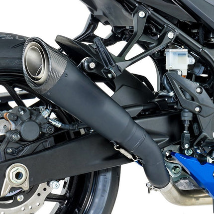 Close-up of a motorcycle exhaust system with a black muffler and a carbon-fiber-textured circular end cap, mounted near the rear wheel on a black bike frame with blue accents.