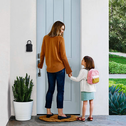 A woman and a child stand at a light blue front door, holding hands. The woman wears a rust-orange sweater and dark jeans, while the child wears a white top, a mint skirt, and a pink and yellow backpack.