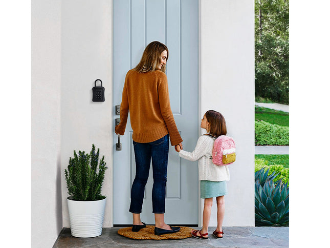 A woman and a child stand at a light blue front door, holding hands. The woman wears a rust-orange sweater and dark jeans, while the child wears a white top, a mint skirt, and a pink and yellow backpack.