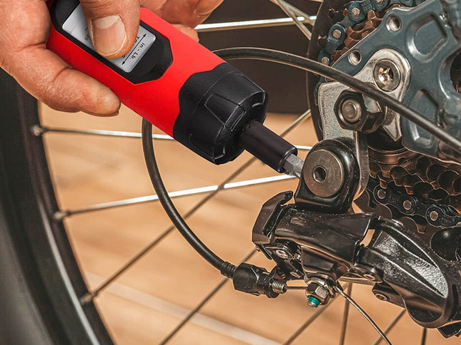A close-up of a person using a red and black handheld torque wrench to tighten a bolt on a bicycle wheel's rear derailleur.