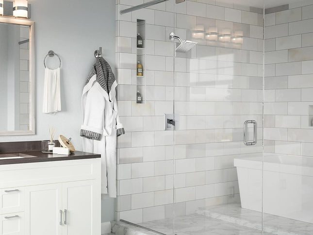 A modern bathroom interior featuring a walk-in glass shower enclosure with white subway tile walls, a white vanity with a dark countertop, a mirror, and hanging white robes.