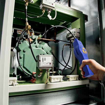 A hand holding a blue handheld diagnostic tool with a yellow logo, pointing it toward the internal components of a large green industrial machine.