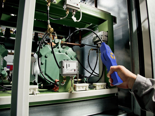 A hand holding a blue handheld diagnostic tool with a yellow logo, pointing it toward the internal components of a large green industrial machine.