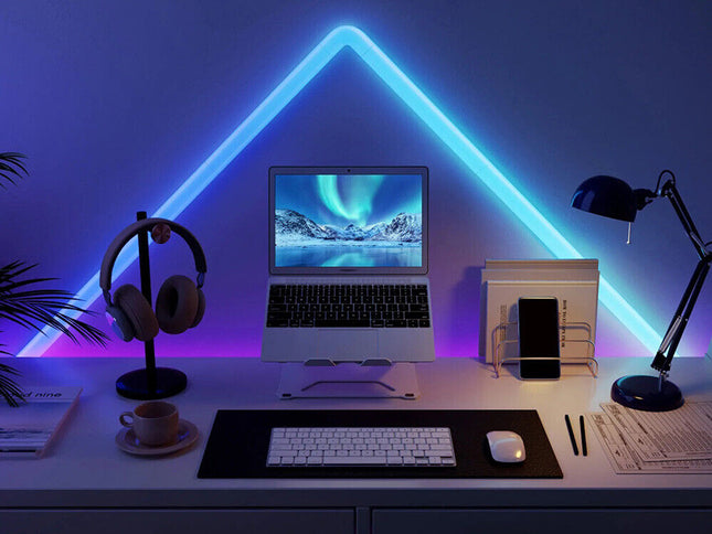 A styled home office desk setup under a glowing blue LED triangle light. The desk features a silver laptop on a stand, a white keyboard and mouse on a black mat, over-ear headphones on a stand, a smartphone in a holder, and a black desk lamp, all illuminated by the blue and purple ambient lighting.