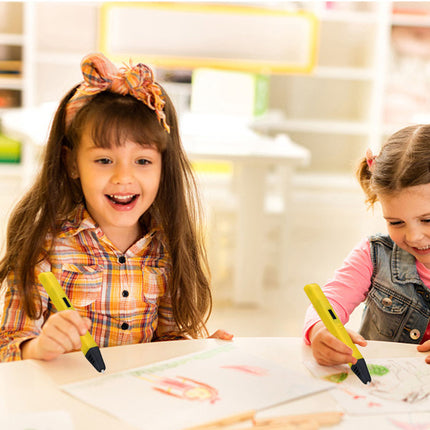 Two young girls smiling and drawing at a table, each holding a yellow 3D pen-style drawing tool in a bright indoor room.