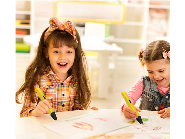 Two young girls smiling and drawing at a table, each holding a yellow 3D pen-style drawing tool in a bright indoor room.
