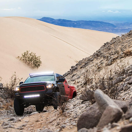 Red off-road truck navigating a rocky desert trail with sand dunes and mountains in the background.