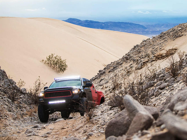 Red off-road truck navigating a rocky desert trail with sand dunes and mountains in the background.