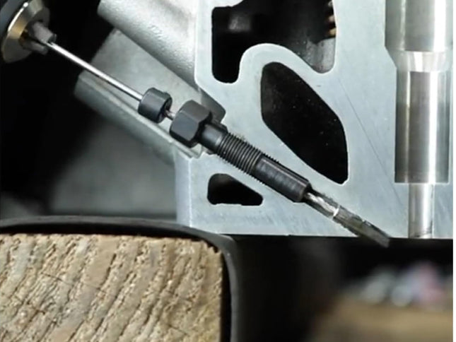 Close-up of a metal mechanical component with a threaded rod or pin and a small metal tool tip, set against a dark industrial background.