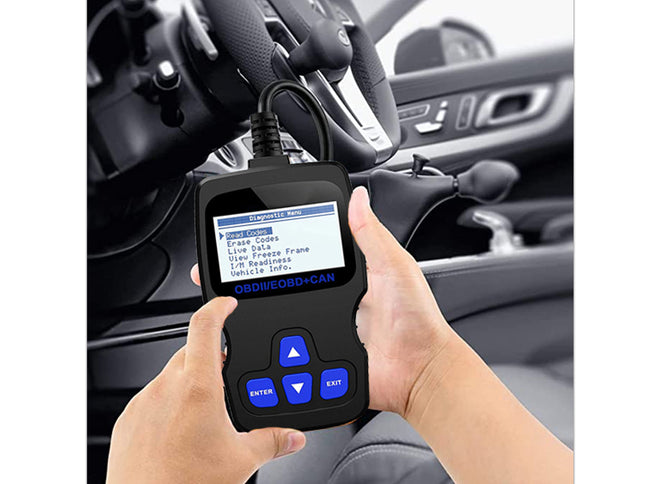 A handheld black OBD2 diagnostic scanner with blue buttons and a screen showing menu options, held by two hands in front of a car dashboard and steering wheel.