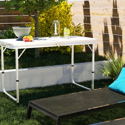White folding table with fruit and glasses on a grassy area with a lounge chair and blue pillow.