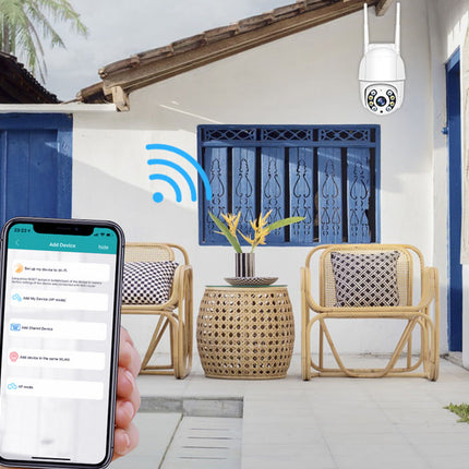A hand holds a smartphone showing a security app interface in the foreground, while a white security camera hangs outdoors on a porch with wicker chairs, blue doors, and a visible wireless signal graphic.