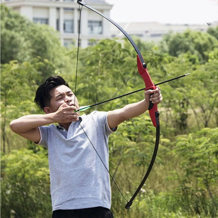 Man in a gray shirt aiming a bow and arrow outdoors with greenery in the background