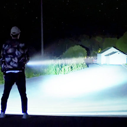 Person using a flashlight on a road at night with buildings and trees in the background