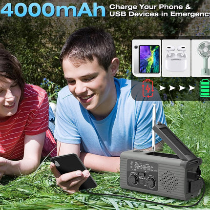 A portable gray emergency radio with an open antenna sits on grass outdoors, with two children lying nearby and a graphic showing it charging a phone, earbuds, and a small fan. Large text reads '4000mAh Charge Your Phone & USB Devices In Emergency'.