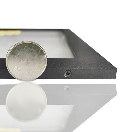 A close-up of a black metal picture frame with a clear glass or acrylic panel, showing a silver coin resting on the left side of the frame. The frame is photographed on a reflective white surface against a white background.