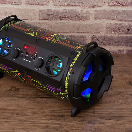 A black cylindrical portable speaker with colorful graffiti-style graphics and glowing blue LED lights, placed on a wooden surface against a brick wall with a laptop partially visible on the right.