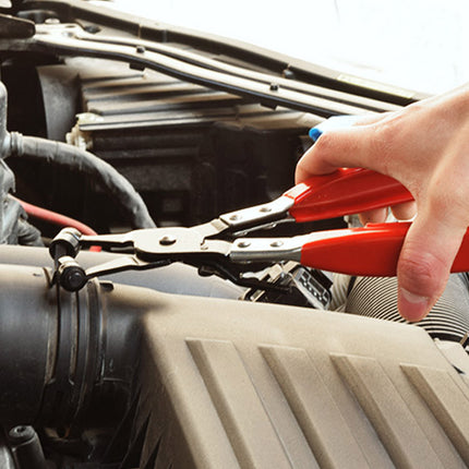 A close-up of a person's hand using red-handled engine pliers to loosen a bolt on a car engine bay.