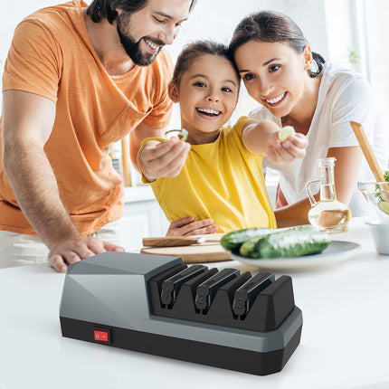 A 4-slice kitchen knife sharpener in black and gray with a red power switch, shown in the foreground with a family happily sharpening vegetables at a kitchen table in the background.