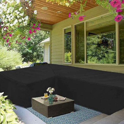 A black outdoor sectional sofa cover fitted over an L-shaped patio sofa on a covered porch, with a small woven coffee table in front and hanging flowers around the house.