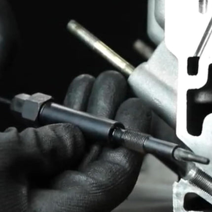 A close-up of a person wearing black gloves holding a black metal alignment tool with a hex nut and screwdriver bit, positioned near a silver metal engine or mechanical component.