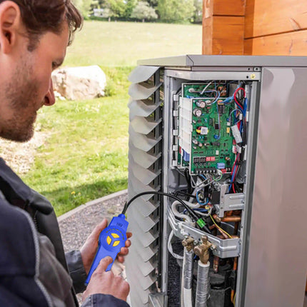 A man in a dark jacket stands beside an outdoor electric heat pump or air conditioner with its side panel open, holding a blue handheld remote control. The unit shows internal wiring and a green circuit board, set next to a wooden wall with grass in the background.