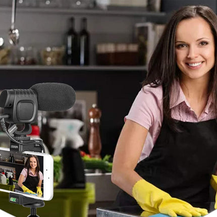 Woman in a kitchen wearing an apron and gloves, with a smartphone camera setup on a stand.
