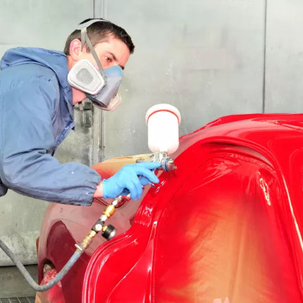 A person in a blue painter's suit and respirator mask uses a spray gun to apply red paint to a vehicle inside a spray booth.