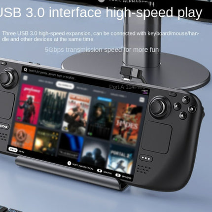 A black handheld gaming console with a screen displaying game thumbnails, shown on a stand on a light blue desk. A monitor stand is behind it, and text at the top reads 'USB 3.