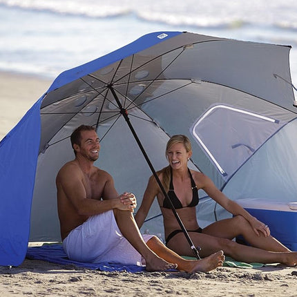 Two people sitting under a large beach umbrella on a sandy beach with a cooler in the background.