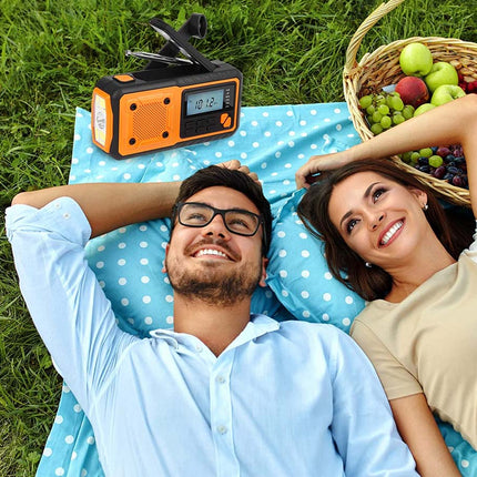 An orange and black portable radio with a clip on top lies on a blue-and-white polka dot blanket on grass, next to a picnic basket of fruit, with a man and woman lying down smiling.