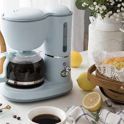 A light blue retro-style drip coffee maker with a glass carafe filled with black coffee, styled on a white table with lemons, a cup of coffee, and a basket of bread in a bright kitchen setting.