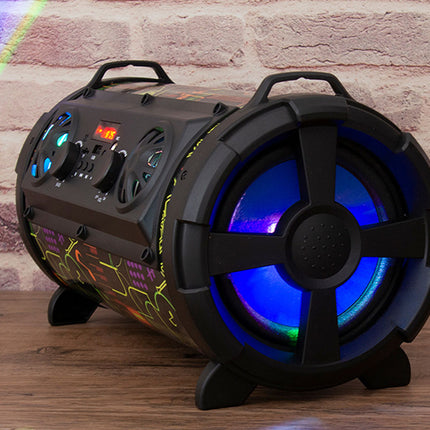 A black portable karaoke speaker with illuminated blue and green LED lights, standing on a wooden surface against a brick wall background.