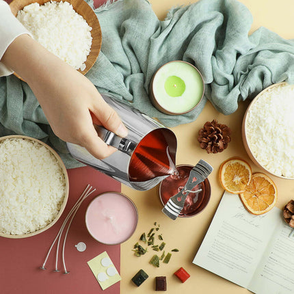 Hand pouring a liquid into a small container surrounded by bowls of rice, a candle, and other items on a textured surface.